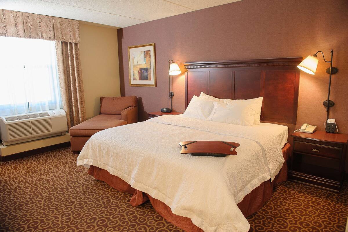 Quarto de hotel com uma cama grande, uma cadeira, uma mesa de cabeceira com abajur, um telefone fixado na parede e uma janela com cortinas.