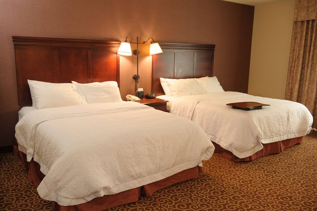 Quarto de hotel com duas camas, uma mesa de cabeceira com abajur e tapete estampado.