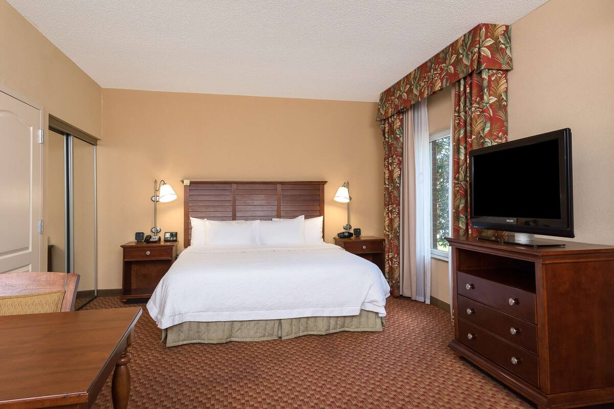Habitación de hotel con una cama grande, dos mesitas de noche con lámparas, un televisor sobre un mueble de madera y una ventana con cortinas.