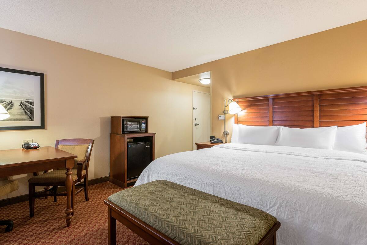 Habitación de hotel con una cama grande, un escritorio con silla, televisión, refrigerador y un cuadro en la pared.