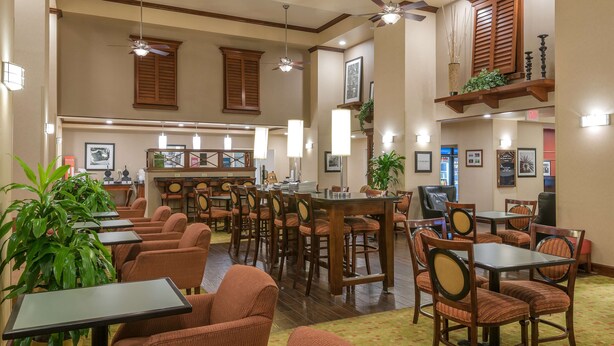 A spacious hotel lobby with multiple seating areas, including tables and chairs, a reception desk, and a staircase leading to an upper level.