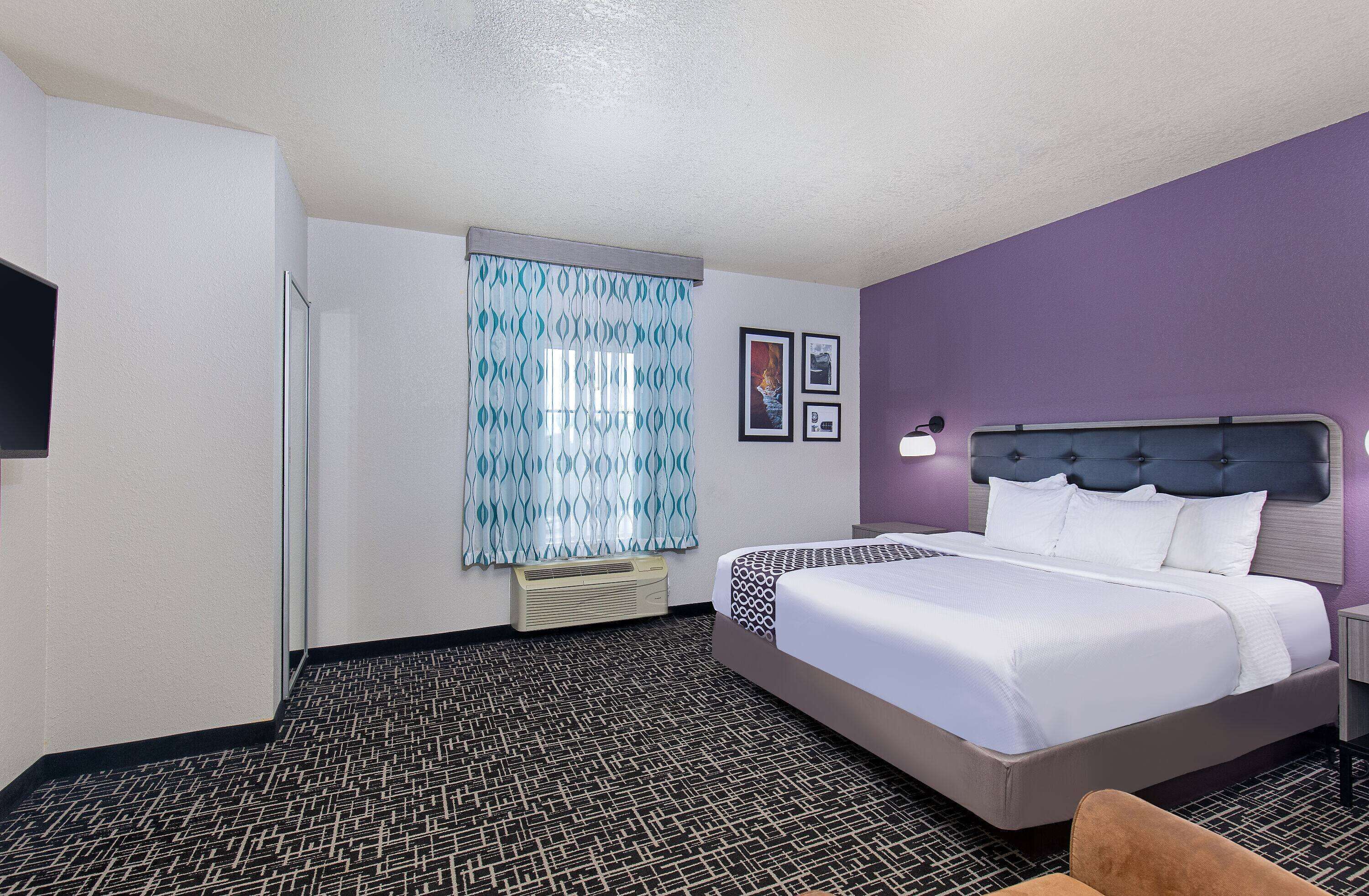 A hotel room with a bed, a nightstand, a lamp, a window with patterned curtains, and a wall-mounted air conditioner.