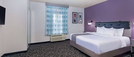 A hotel room with a bed, a nightstand, a lamp, a window with patterned curtains, and a wall-mounted air conditioner.