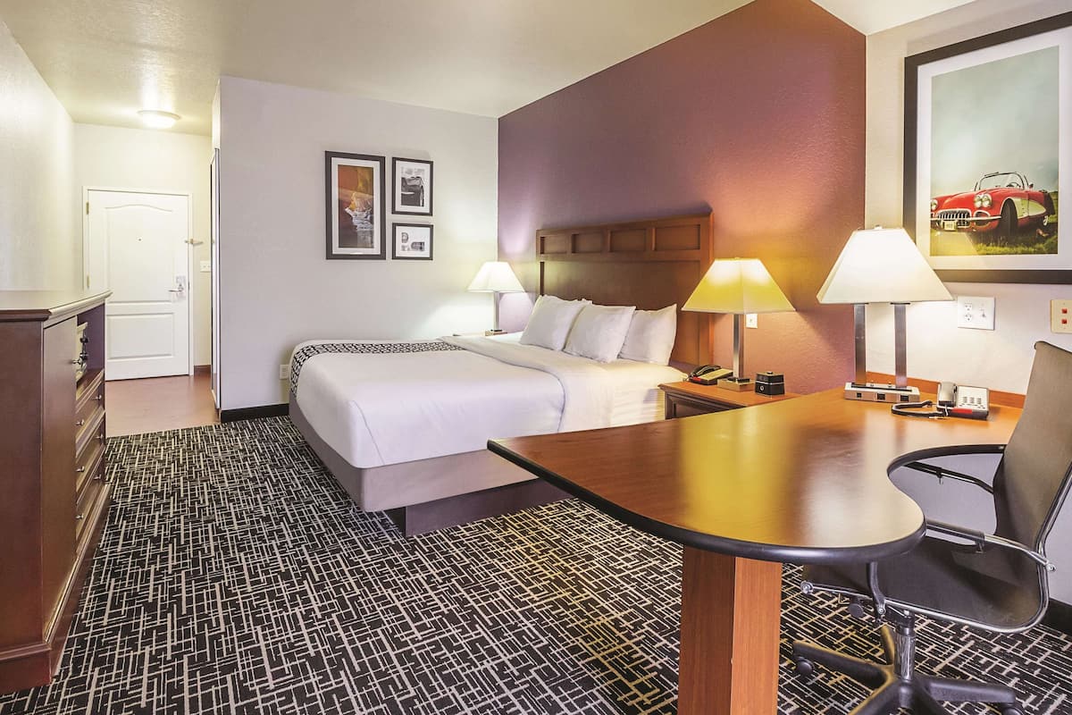 A hotel room with a bed, two bedside tables with lamps, a desk with a chair, a patterned carpet, and a door.