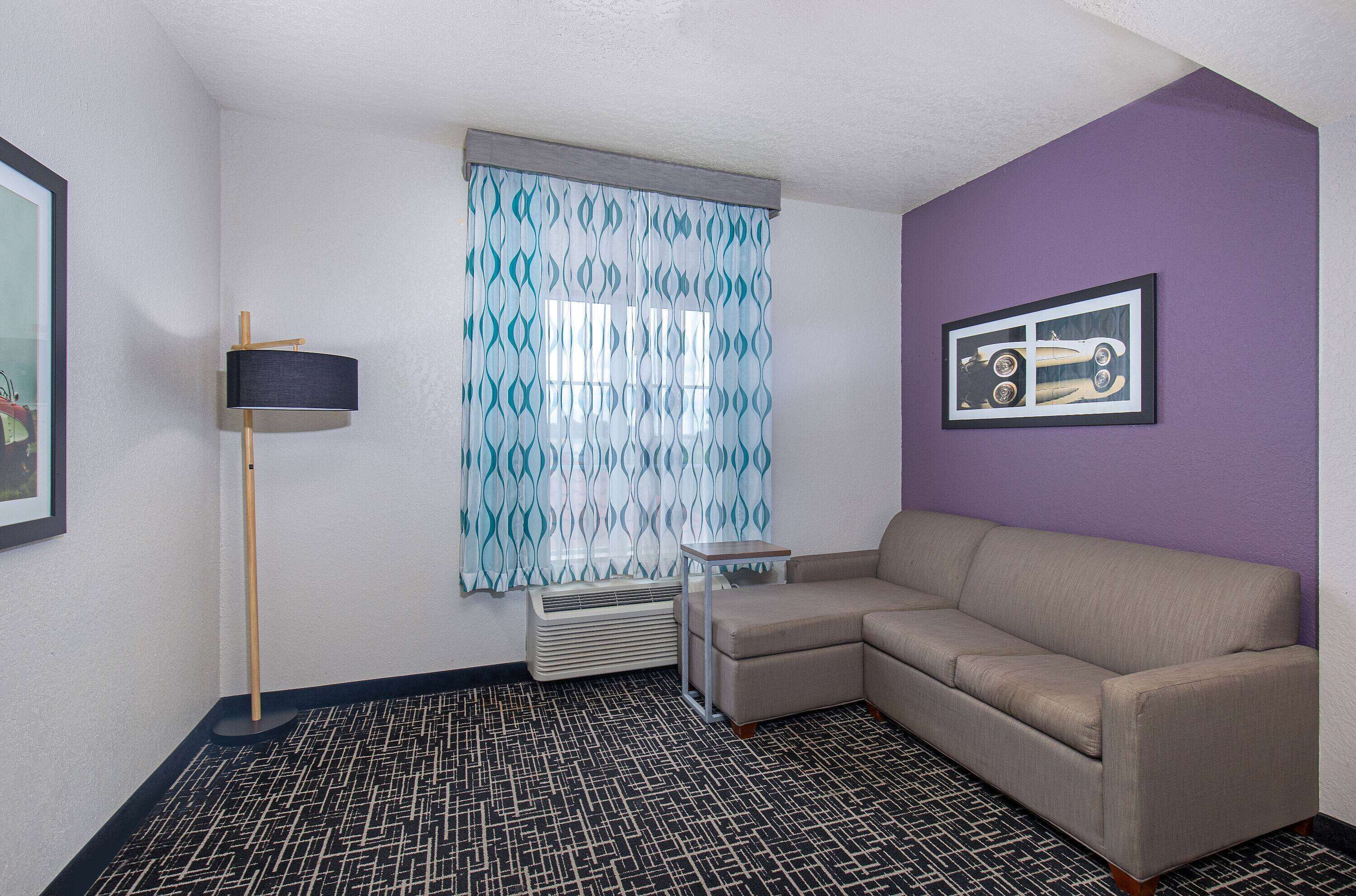 A room with a beige sectional sofa, a standing floor lamp, a patterned floor, a window with blue and white curtains, and a framed picture on the wall.