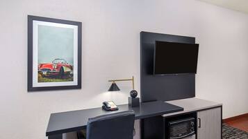A modern hotel room with a desk, a flat-screen TV mounted on the wall, a framed picture of a red car, a desk lamp, and a built-in oven.