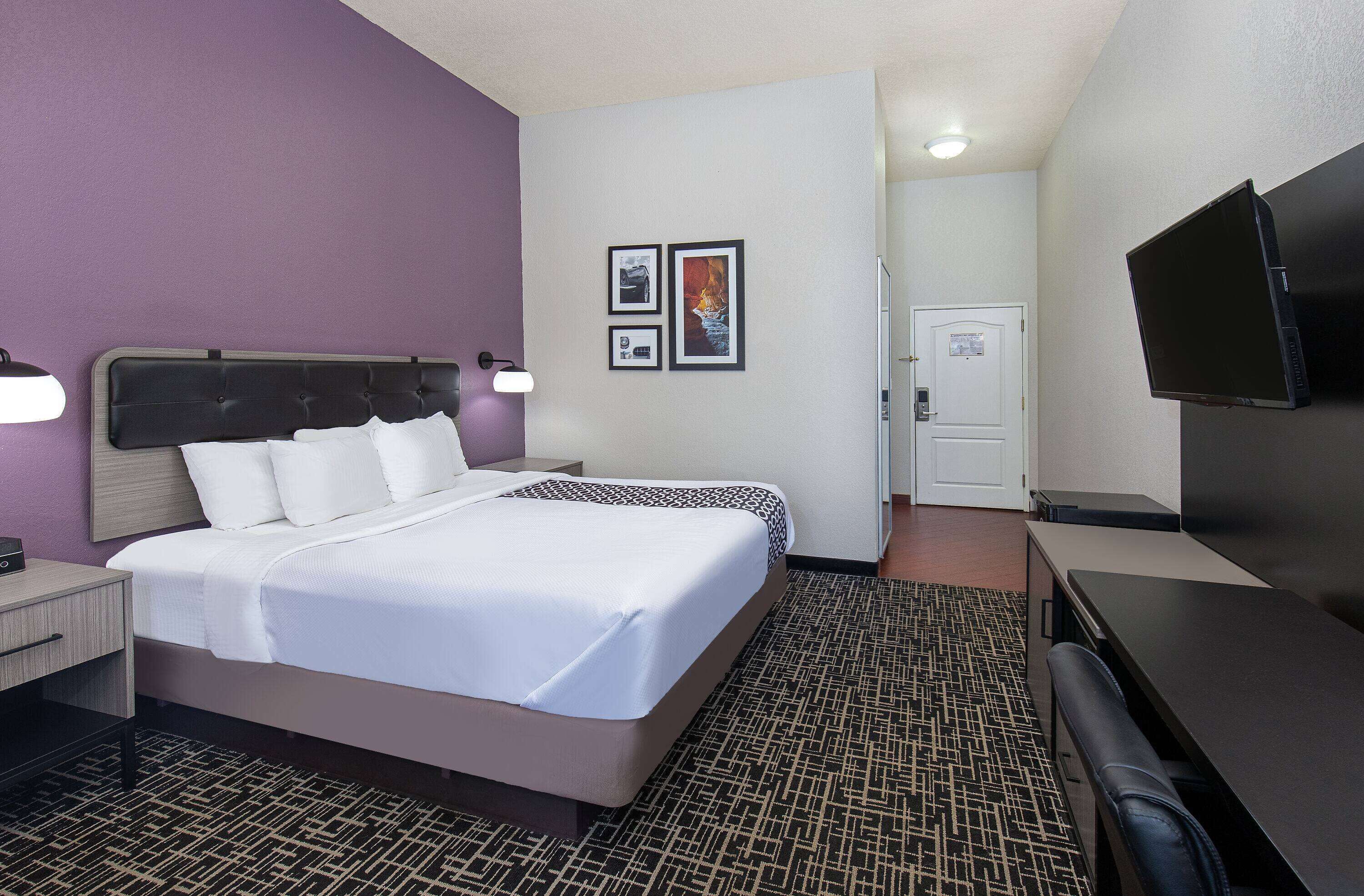 Una habitación de hotel con cama, mesita de noche, lámpara, televisor y una puerta que da a otra habitación.