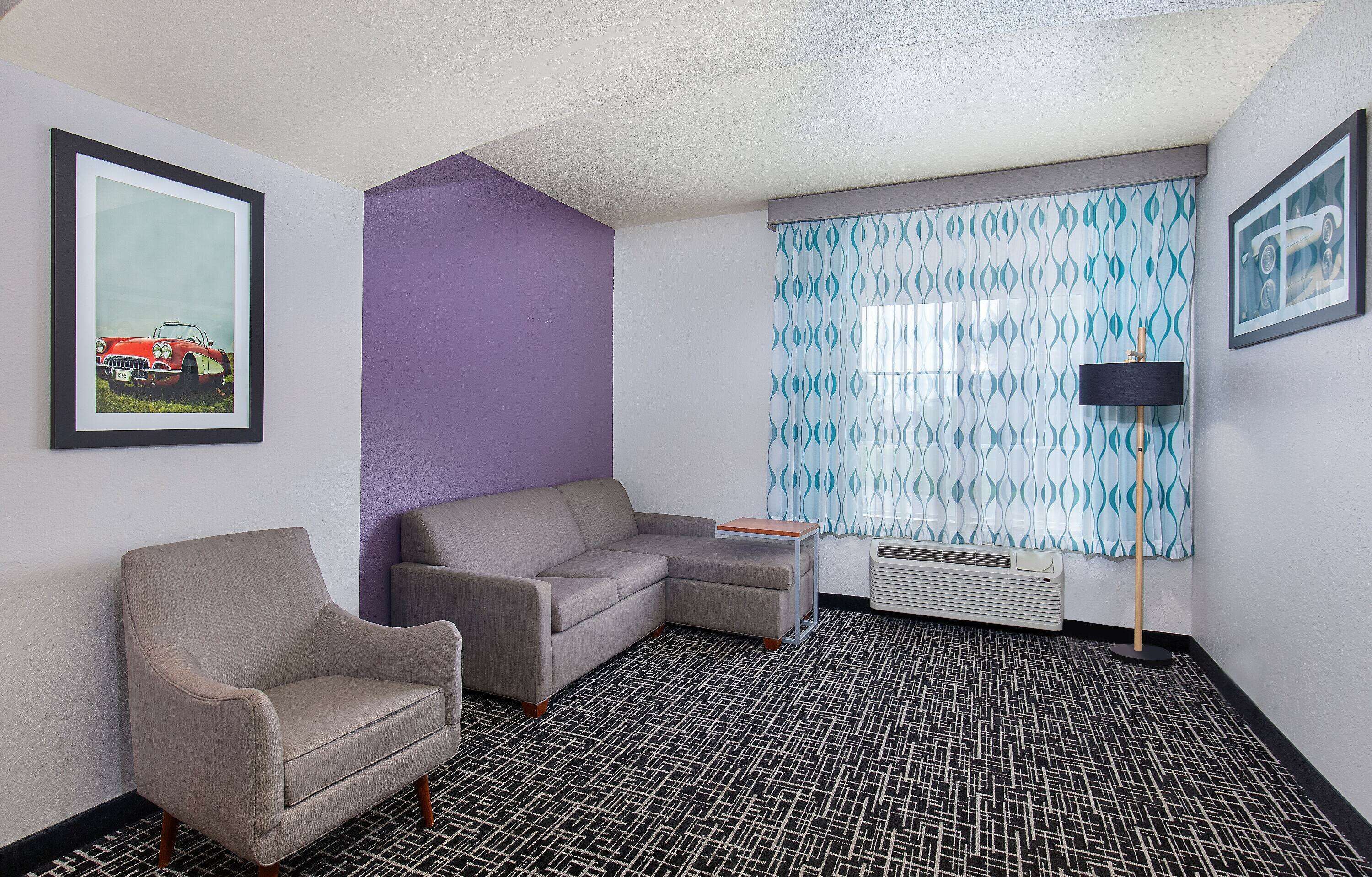 A hotel room with a sectional sofa, a single armchair, a small table, a patterned floor, and a window with patterned curtains.