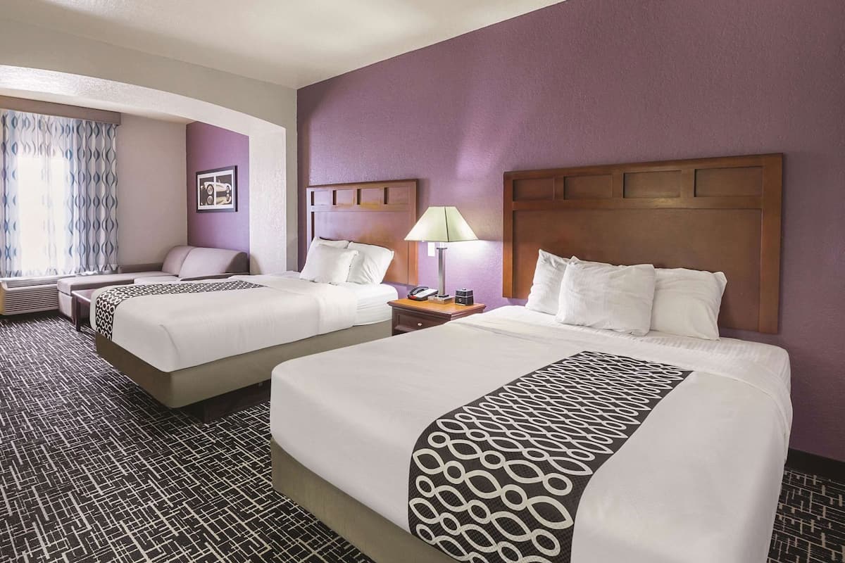 A hotel room with two beds, a bedside table with a lamp, a chair, and a window with curtains.