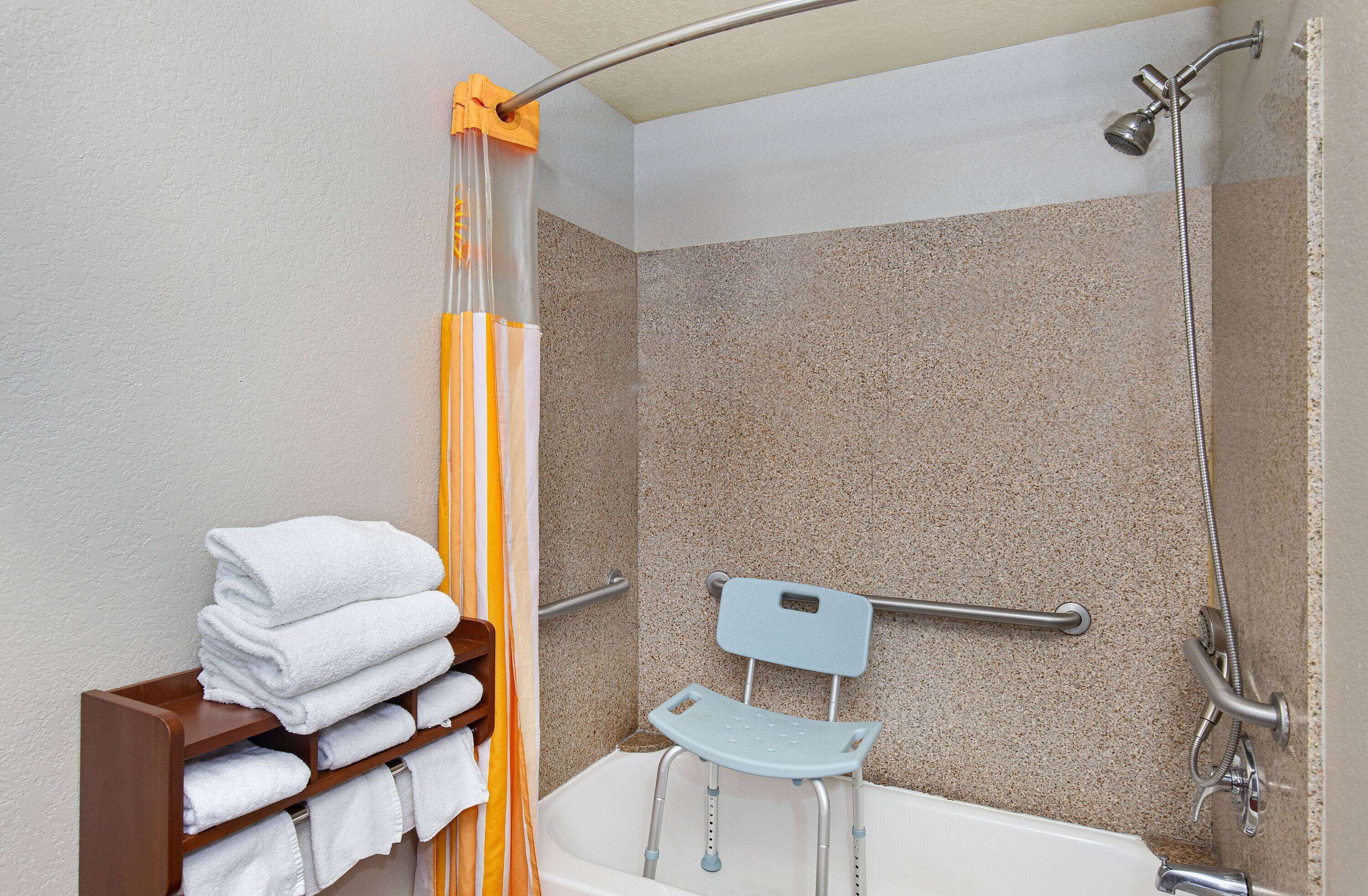 A bathroom with a shower, grab bars, a wheelchair-accessible bench, and a stack of towels on a shelf.