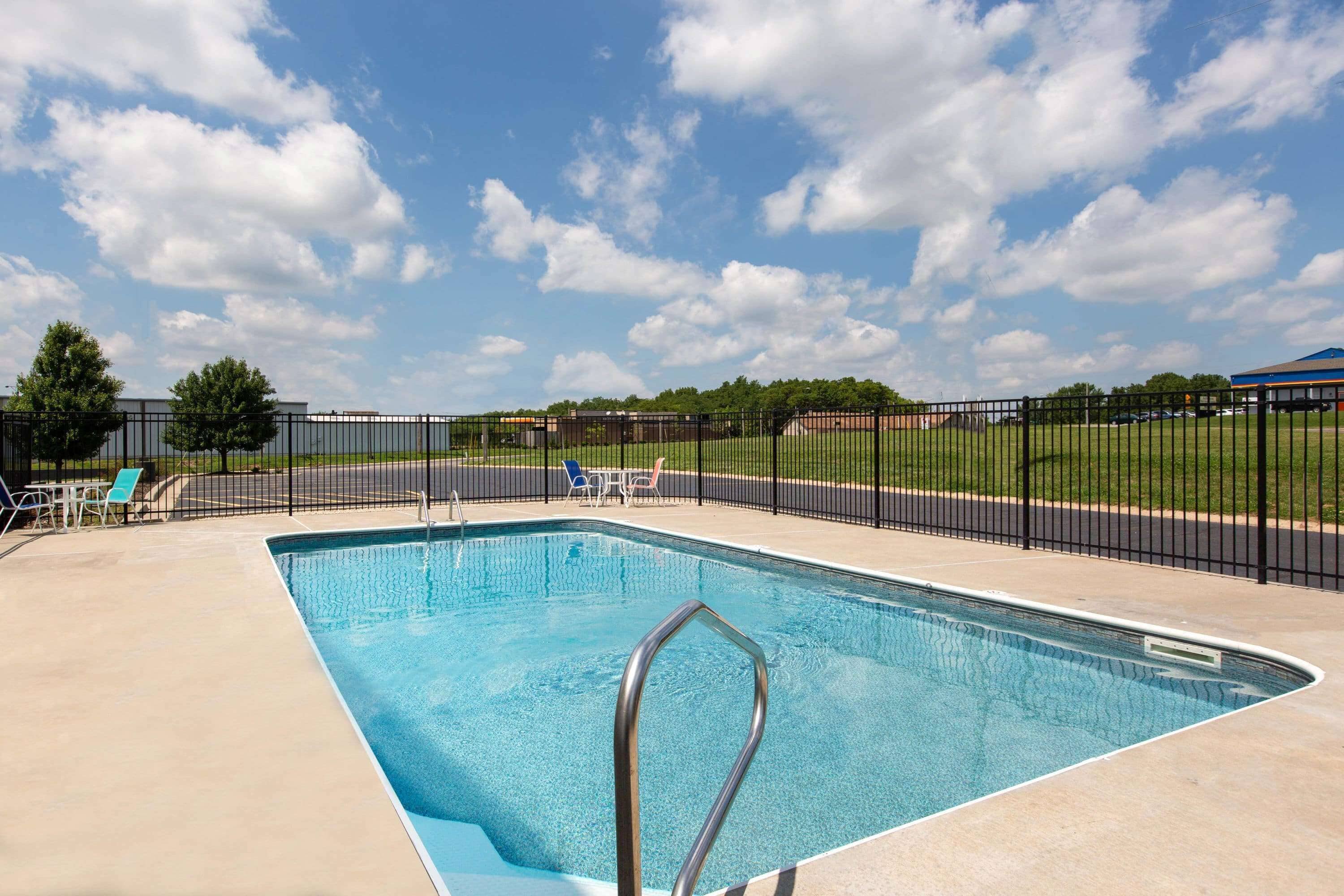A rectangular swimming pool with a metal ladder, surrounded by a black fence, and a clear blue sky with clouds.