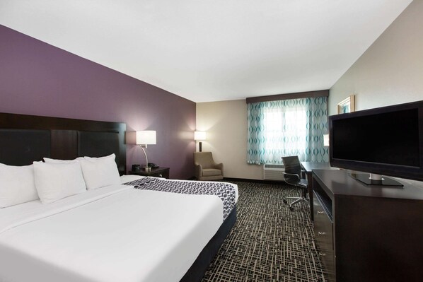 A hotel room with a large bed, a desk with a flat-screen TV, a chair, a lamp, and a window with curtains. - La Quinta Inn & Suites by Wyndham Blue Springs (Blue Springs)