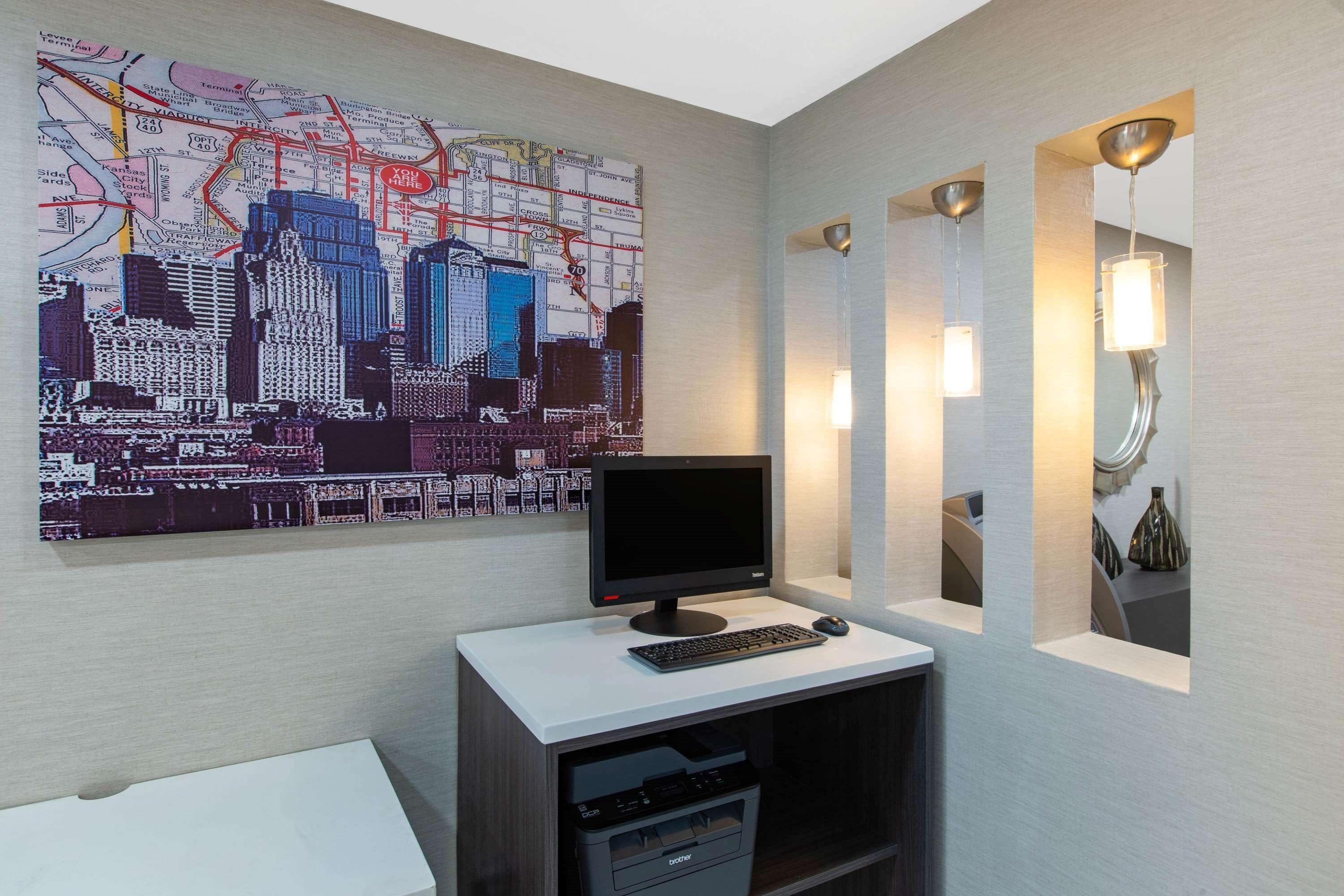 A hotel room with a cityscape artwork on the wall, a desk with a computer, and a mirror with integrated lighting.