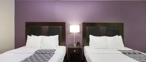 Habitación de hotel con dos camas, una mesita de noche con lámpara y una alfombra estampada.