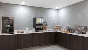 A modern kitchen with a self-service station, including a refrigerator, coffee machines, and a food display.