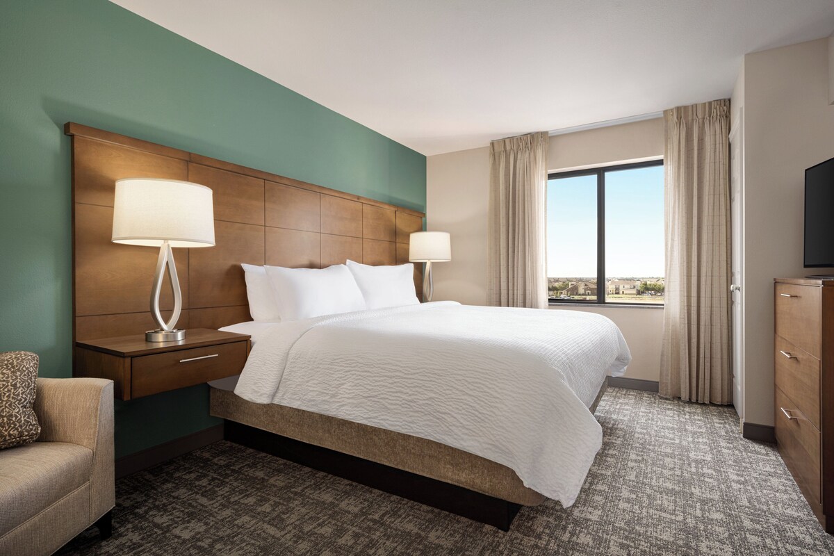 A hotel room with a large bed, two bedside tables with lamps, a TV, a dresser, and a view of the city.