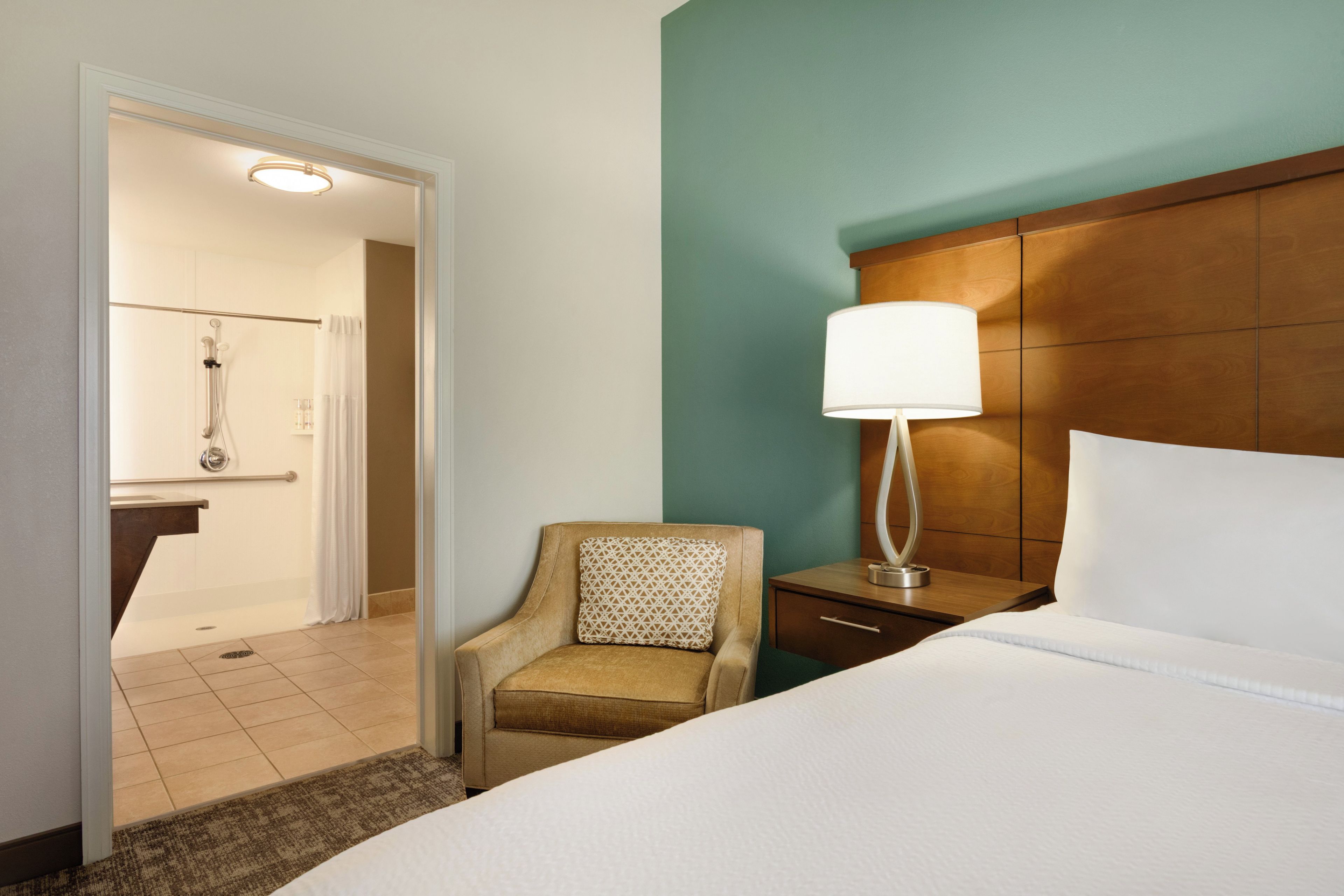 Une chambre d’hôtel avec un lit, une table de chevet avec une lampe, une chaise et une salle de bain visible à travers une porte ouverte.