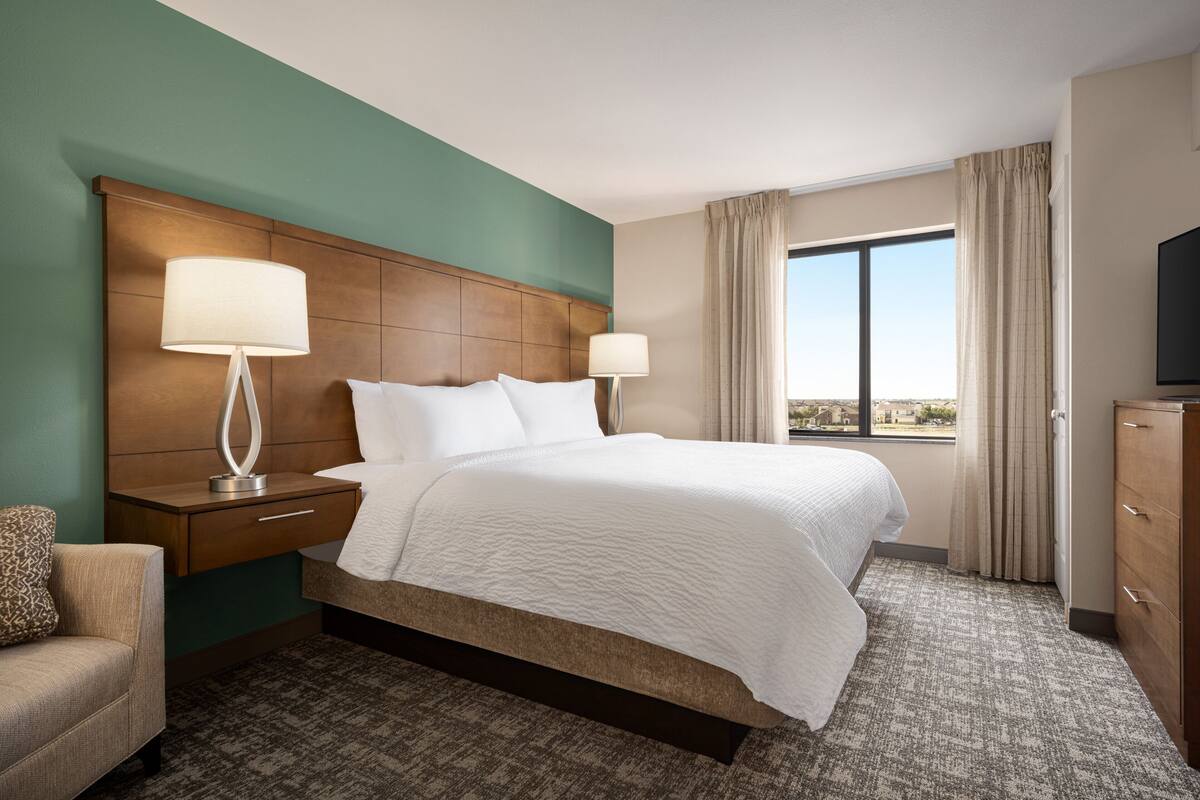 Habitación de hotel con una cama grande, dos mesitas de noche con lámparas, un televisor, un aparador y vistas a la ciudad.