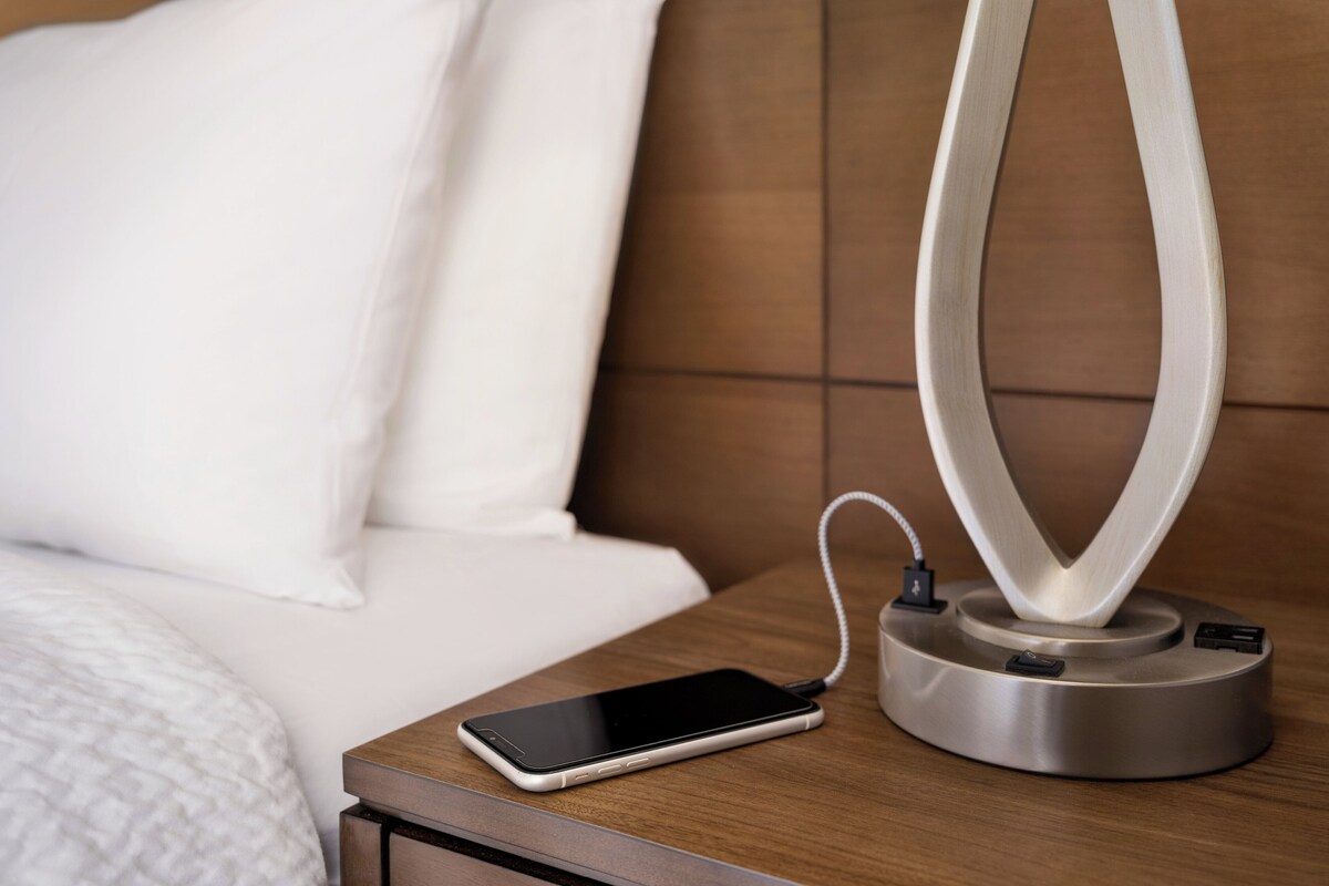 A smartphone on a bedside table with a charging cable, a modern lamp, and a wooden headboard.