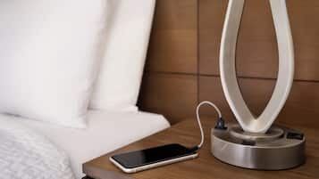 Un teléfono inteligente sobre una mesita de noche, con un cable de carga conectado a una lámpara moderna y de diseño curvo.