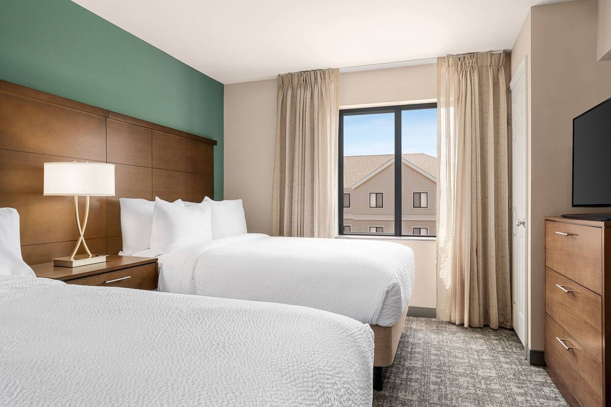 Habitación de hotel con dos camas, cabecera de madera, mesita de noche con lámpara, cómoda y una ventana grande con cortinas.