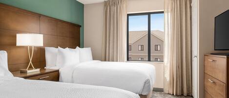 A hotel room with two beds, a wooden headboard, a nightstand with a lamp, a dresser, and a large window with curtains.