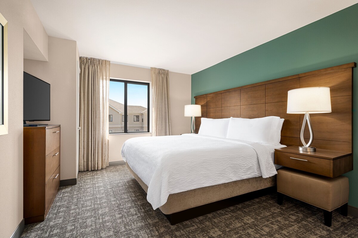Habitación de hotel con una cama grande, cabecera de madera, dos mesitas de noche con lámparas, televisor de pantalla plana, cómoda y vista a edificios a través de una ventana con cortinas.