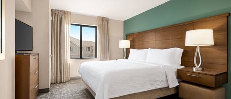 A hotel room with a large bed, a wooden headboard, two bedside tables with lamps, a flat-screen TV, a dresser, and a view of buildings through a window with curtains.