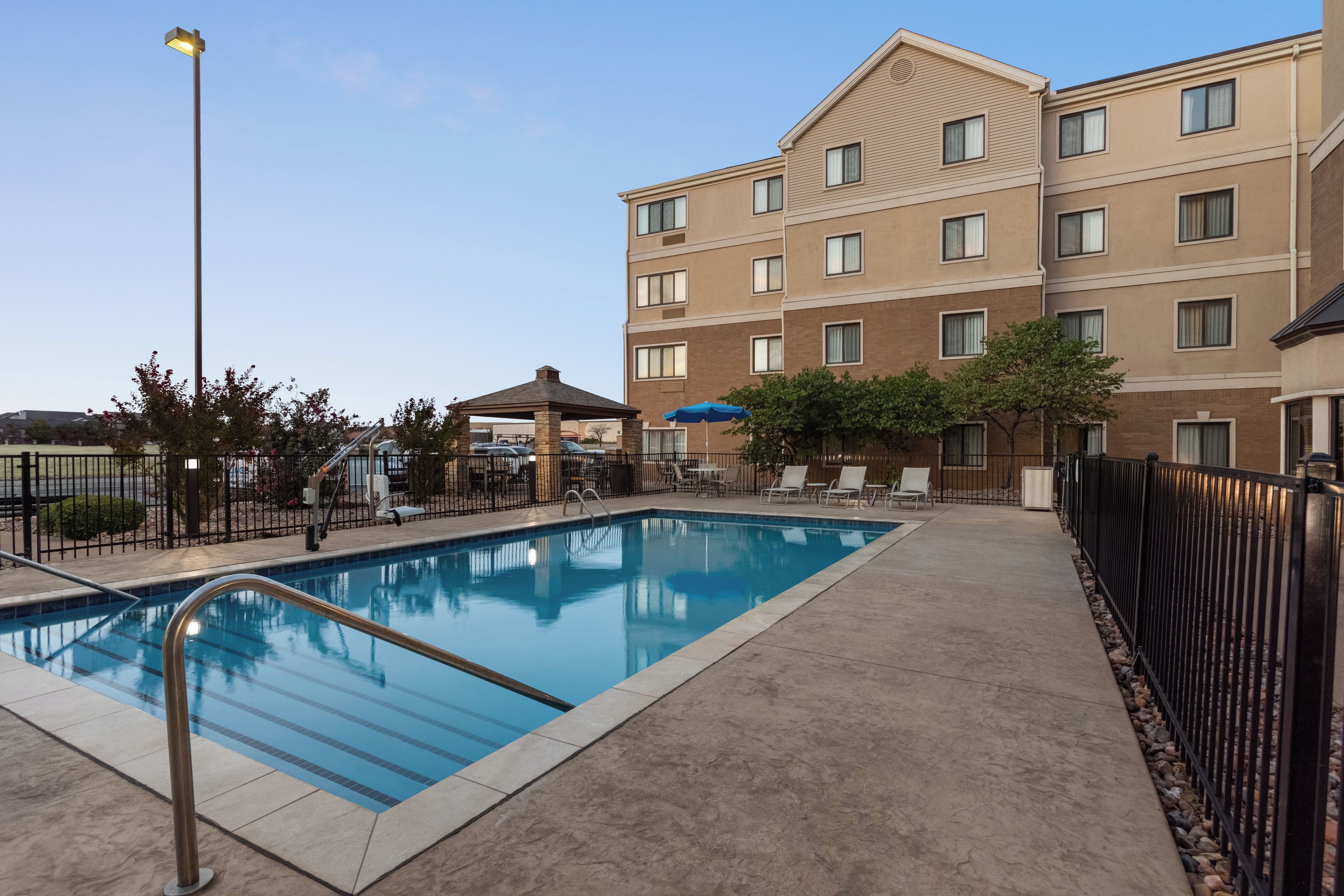 Una piscina con una escalera metálica, rodeada por una terraza de hormigón y una valla. Se observan tumbonas y sombrillas al fondo.