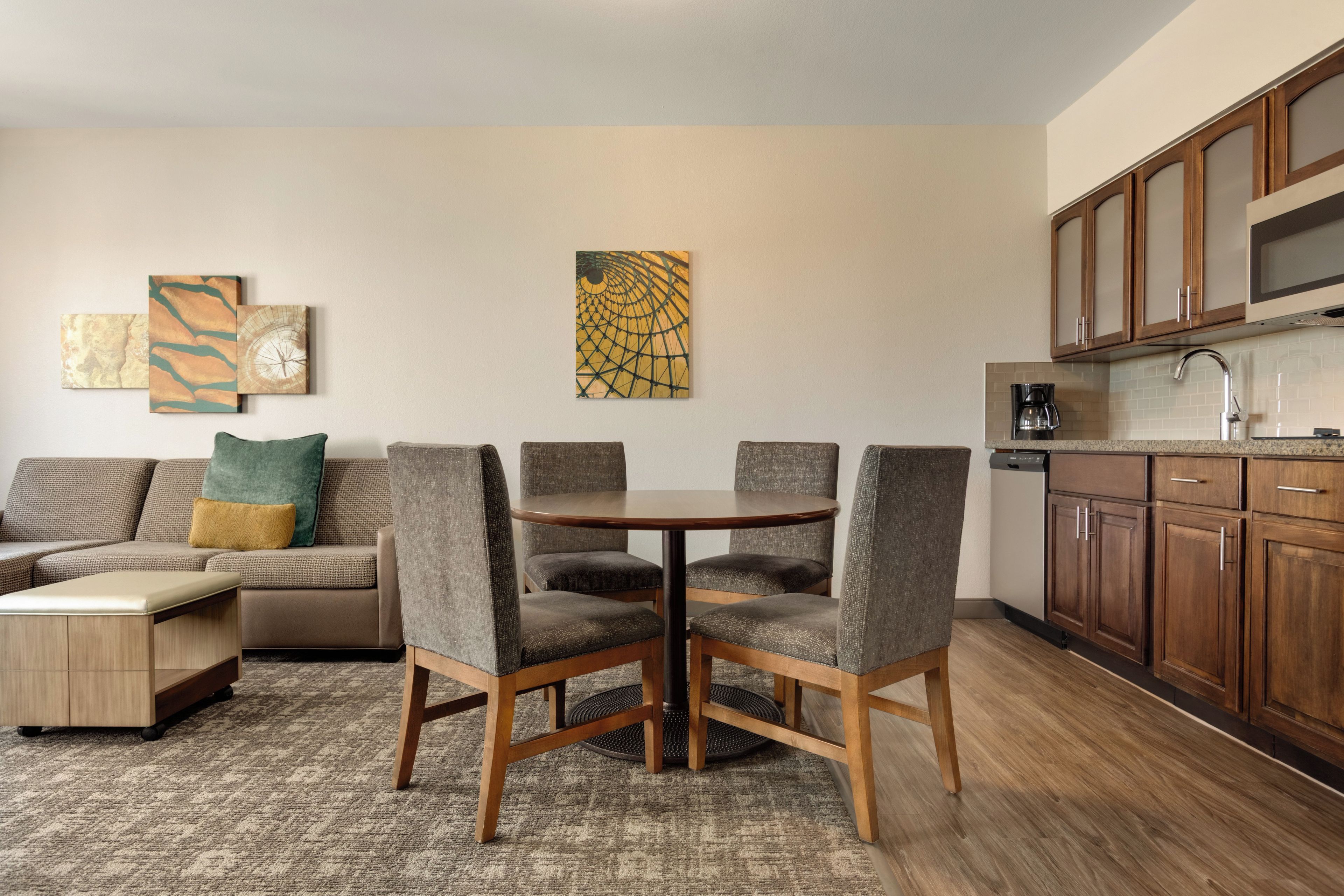 Een moderne woonkamer met een bank, een eettafel en stoelen, een keuken met houten kasten en abstracte muurkunst.