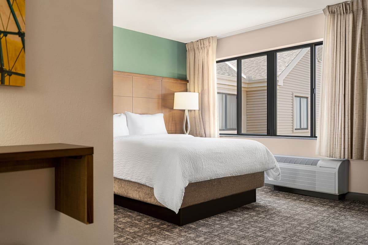 Una habitación de hotel con cama, mesita de noche, lámpara, ventana con cortinas y unidad de aire acondicionado.