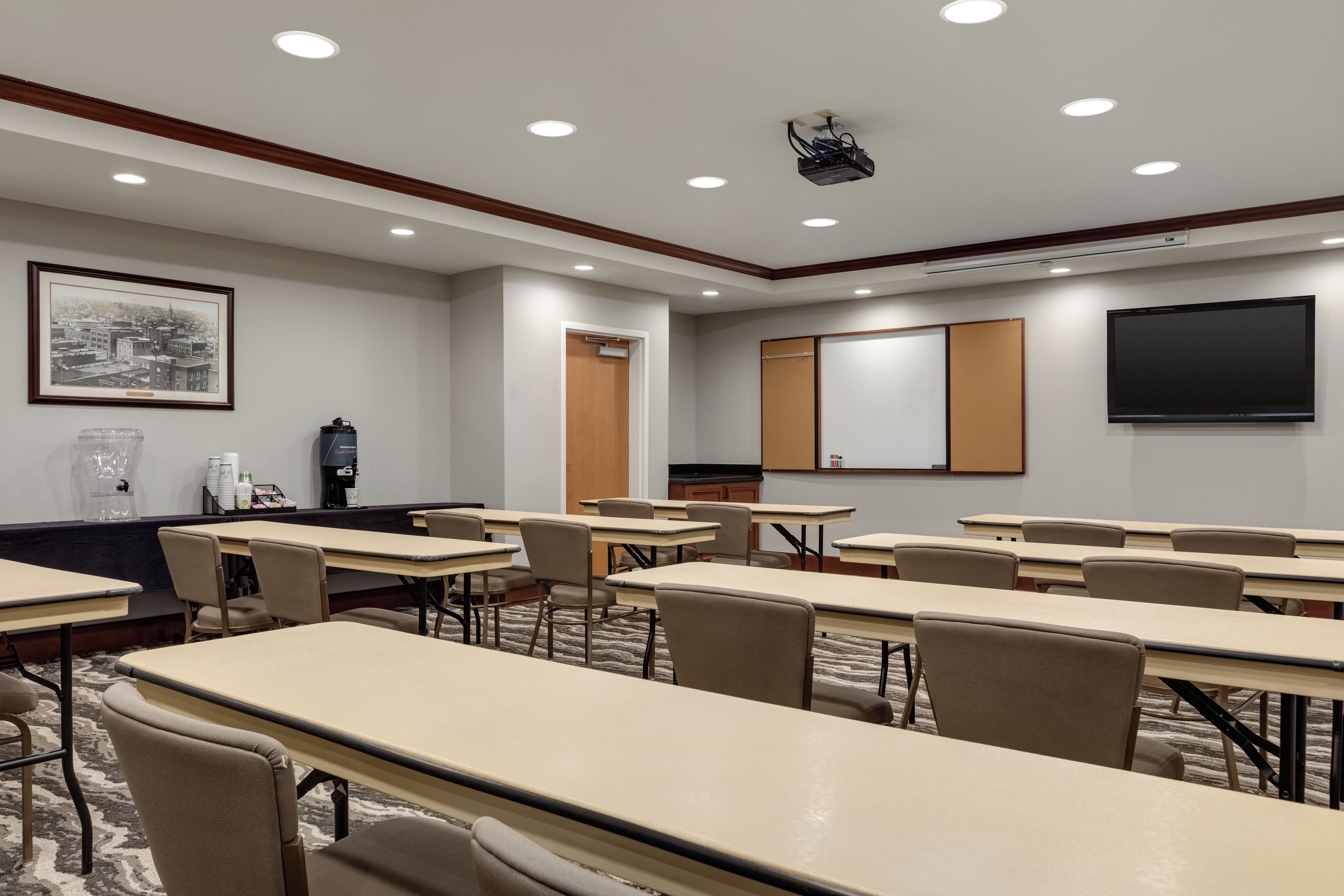 Un salón de conferencias con varias mesas y sillas, un proyector, un cuadro grande en la pared y una estación de café.
