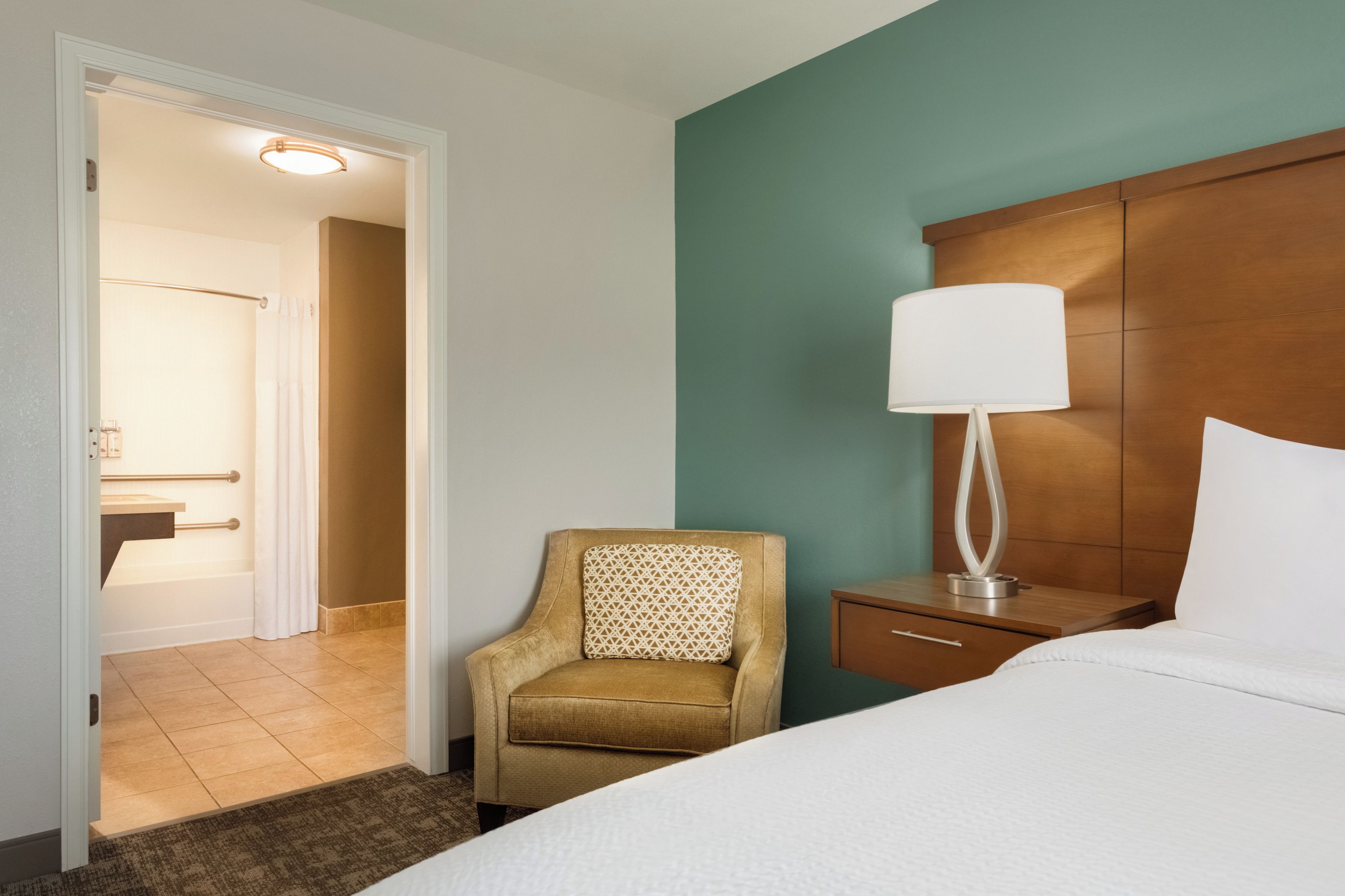 A hotel room with a bed, a nightstand, a lamp, a chair, and a view of the bathroom.