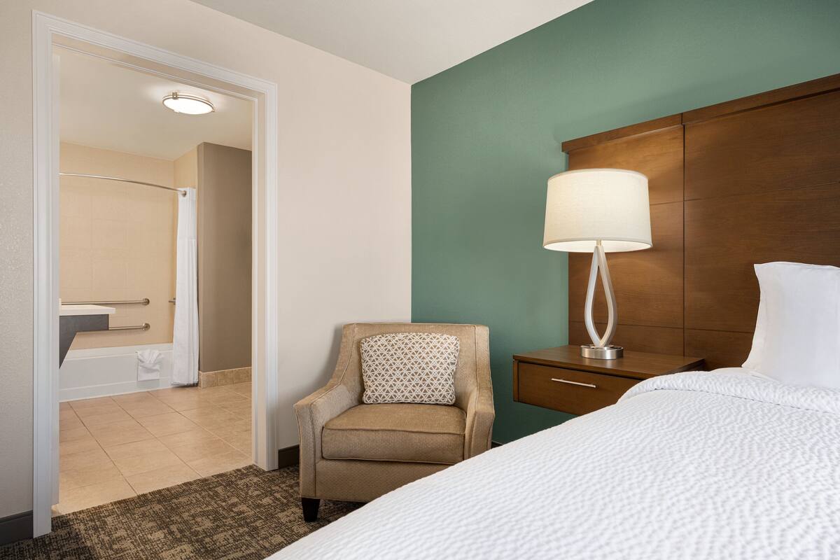 Habitación de hotel con cama, una silla, mesita de noche, lámpara y vista al baño.
