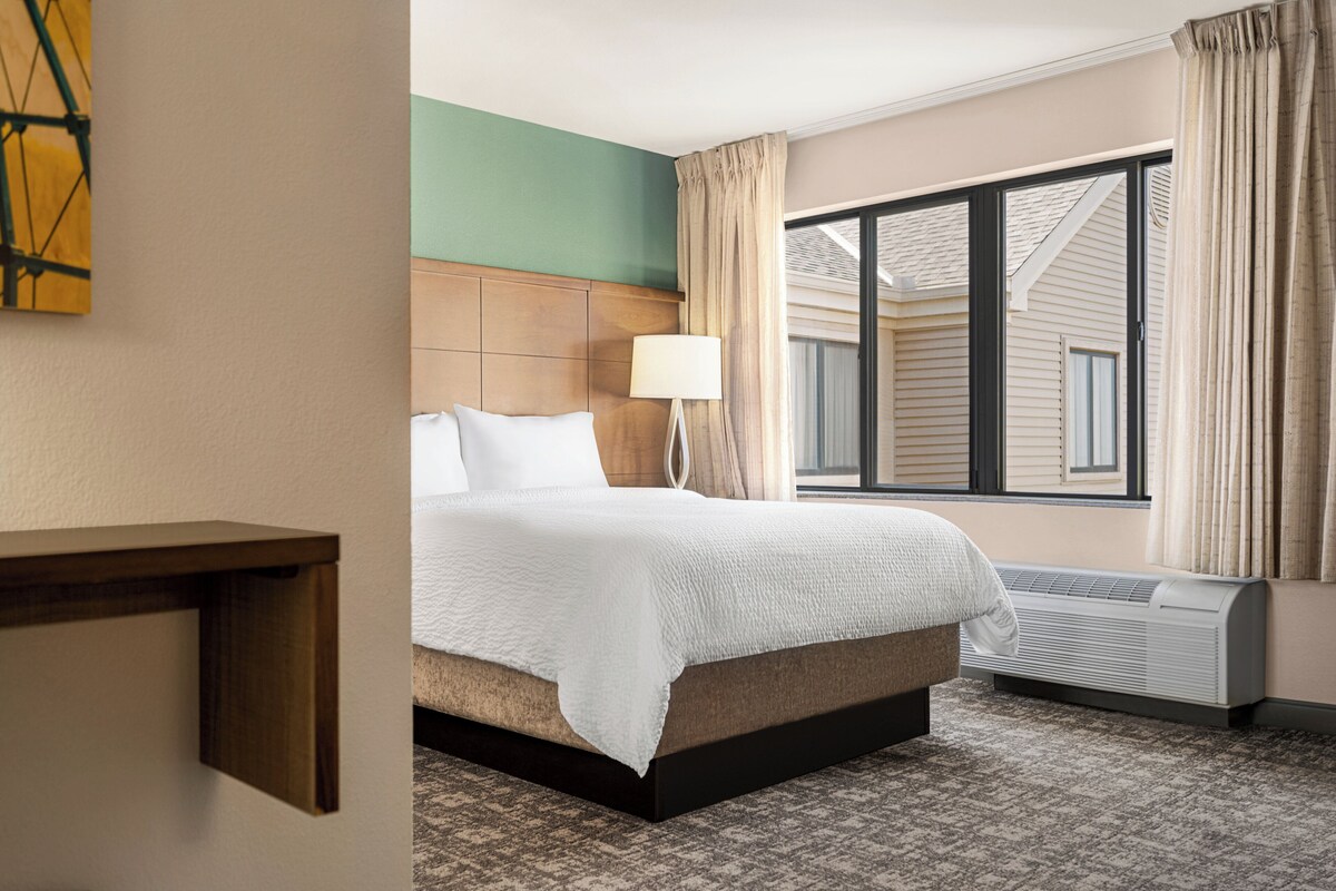A hotel room with a bed, a bedside table, a lamp, a window with curtains, and an air conditioning unit.