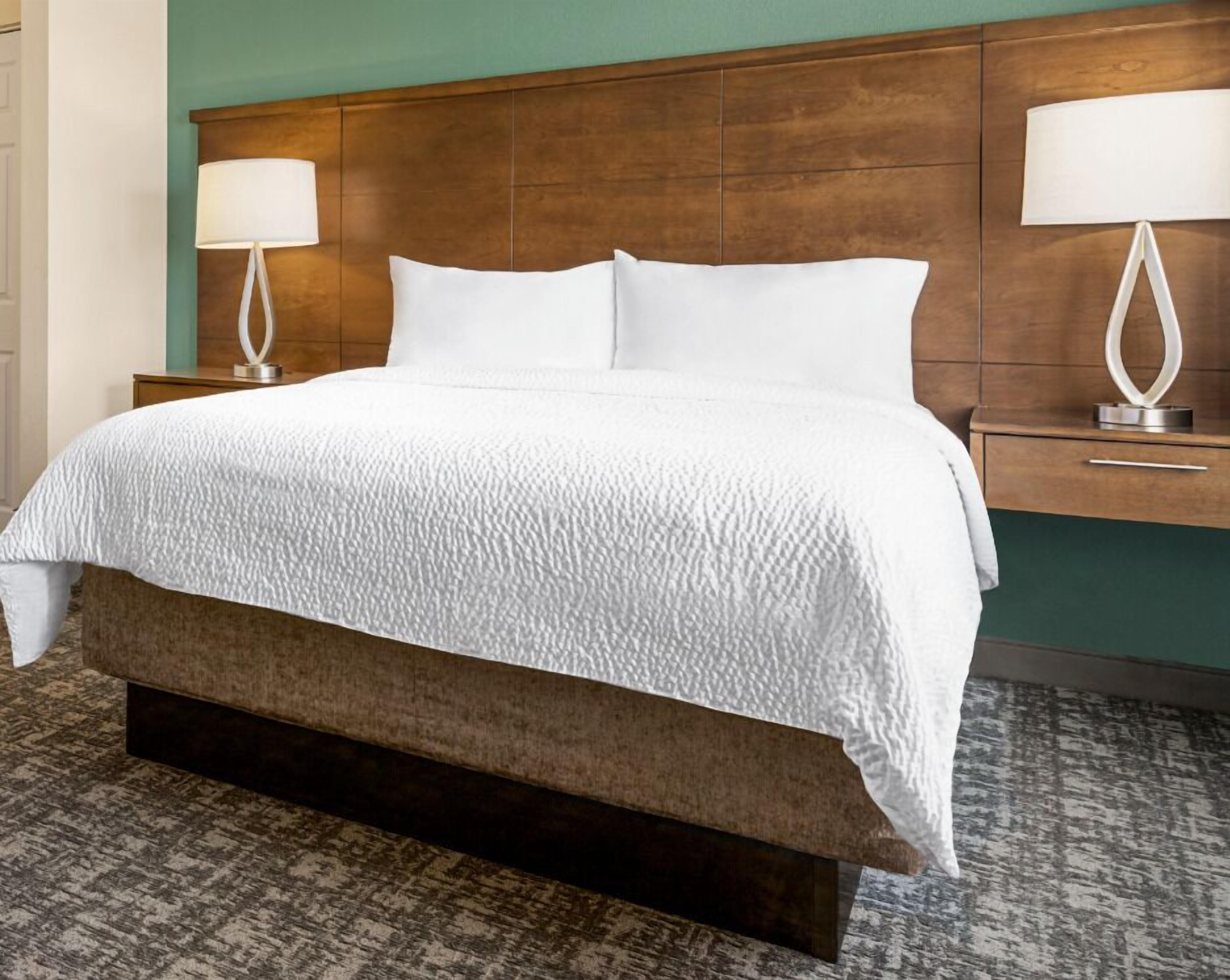 Una habitación de hotel con una cama grande, cabecera de madera, dos mesitas de noche con lámparas y una alfombra estampada.