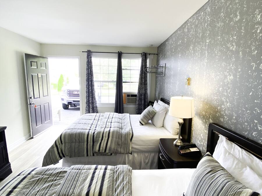 A hotel room with two beds, a nightstand with a lamp, a wall-mounted TV, and a window with curtains.