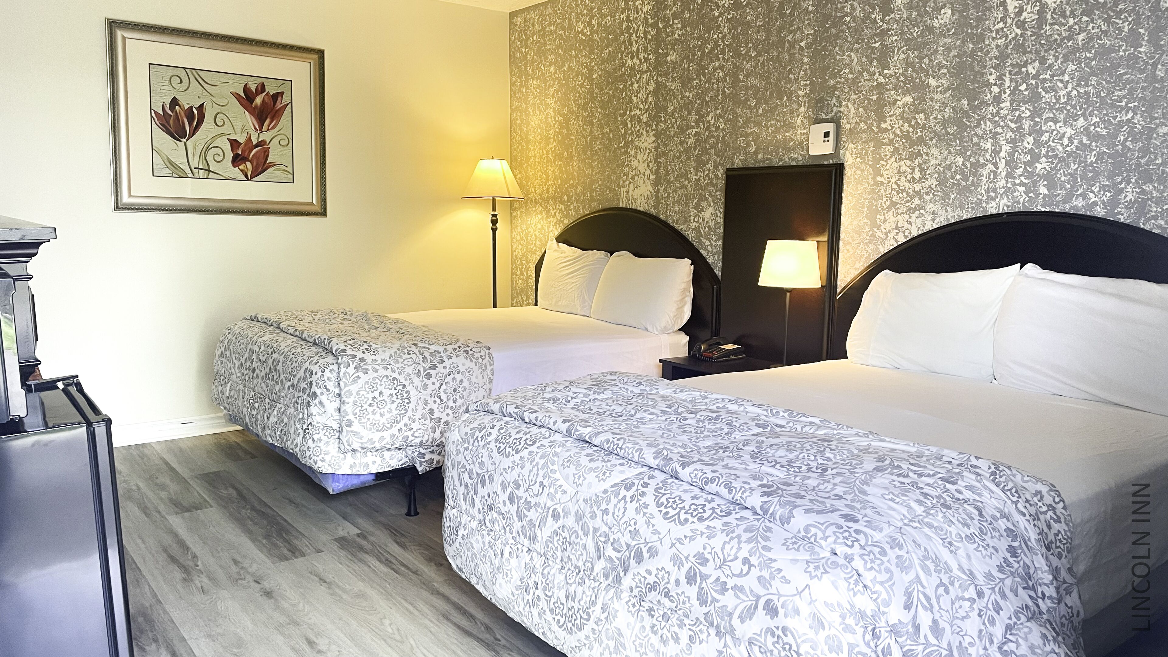 A hotel room with two beds, a nightstand with a lamp, a framed floral artwork on the wall, and a thermostat.