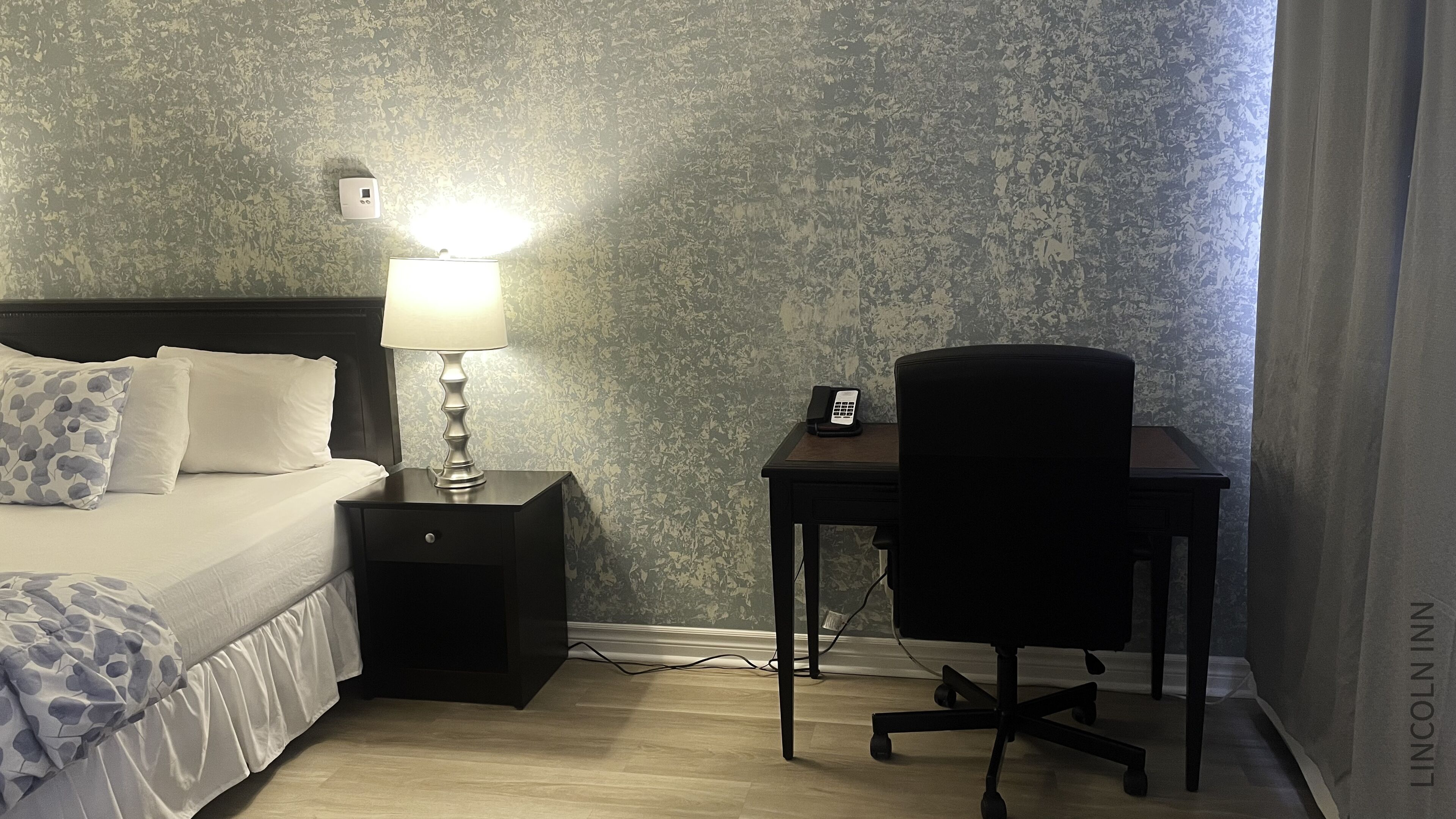 A hotel room with a bed, a nightstand with a lamp, a desk with a phone, and a chair.