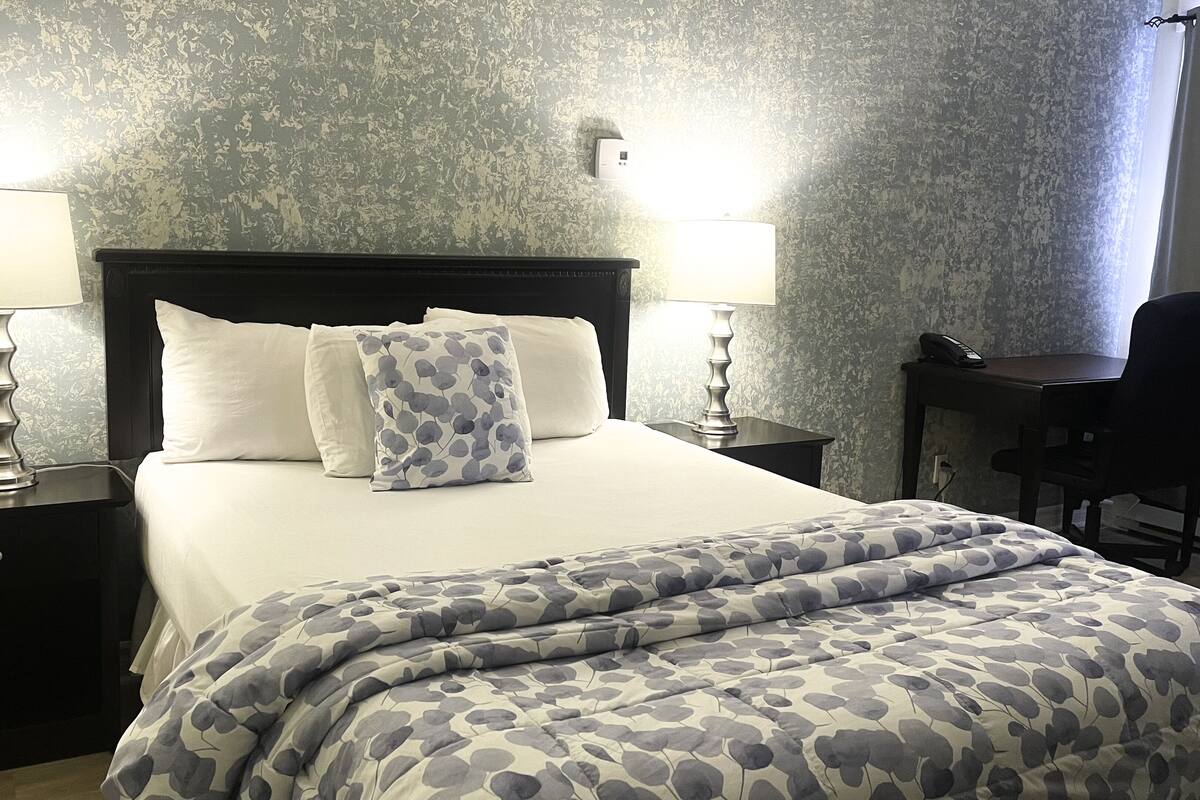 Una cama bien hecha con un edredón estampado y almohadas, dos lámparas de noche, un escritorio pequeño con una silla y una pared con textura.