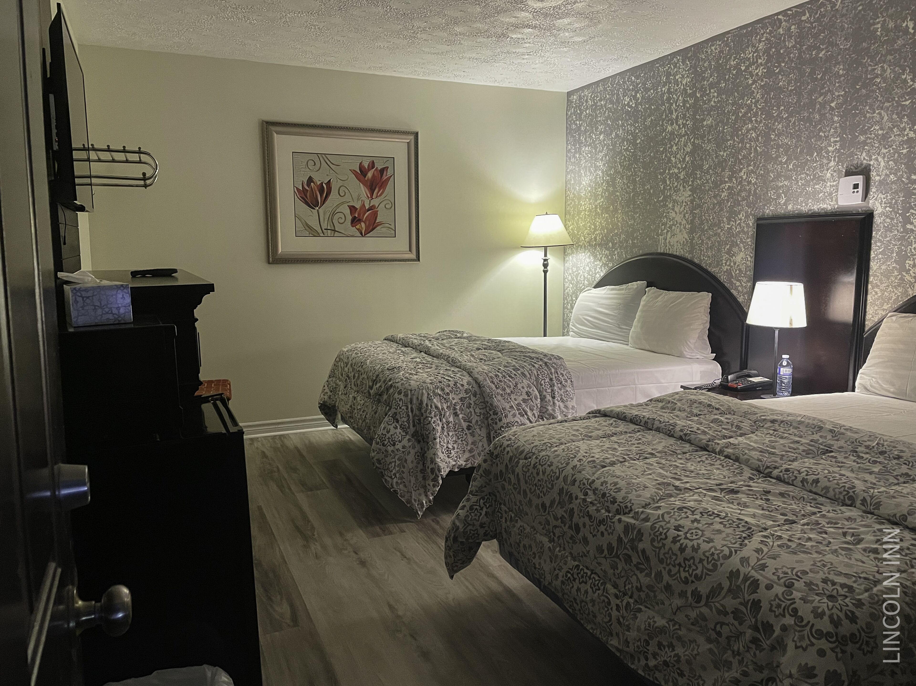 A hotel room with two beds, a nightstand with a lamp, a framed picture on the wall, and a wooden floor.