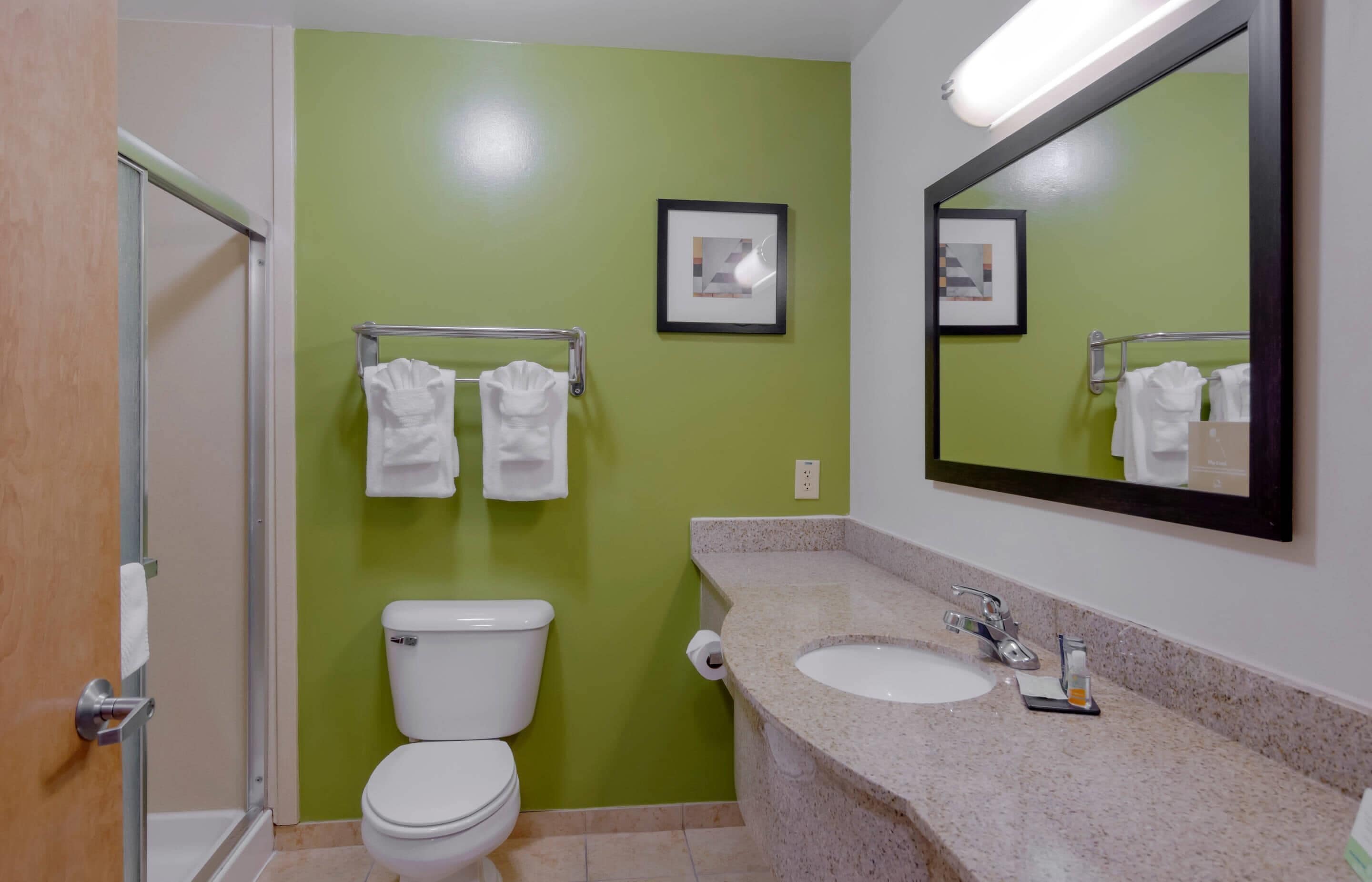 Un baño con pared verde, inodoro blanco, lavamanos con espejo y dos toallas colgadas en un perchero.