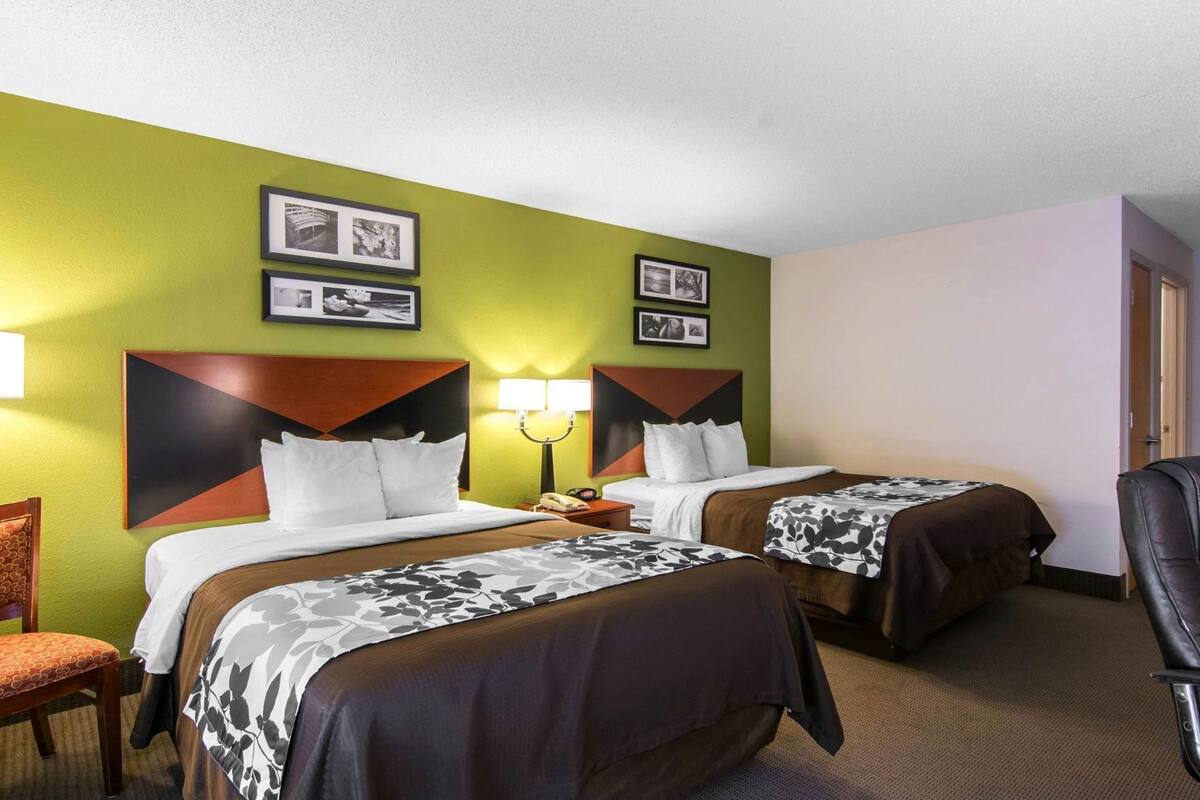 A hotel room with two beds, a desk, a chair, and a lamp.