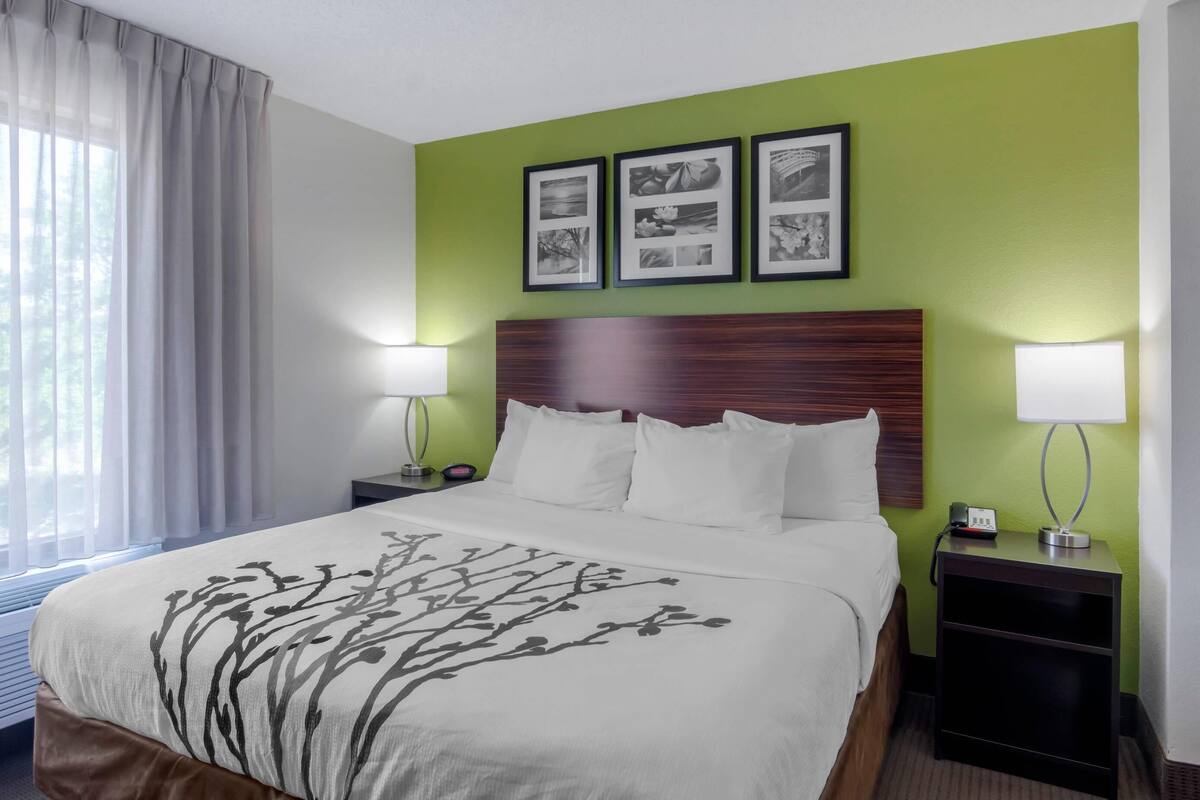A hotel room with a bed, two bedside tables, a nightstand, a lamp, and a window with curtains.