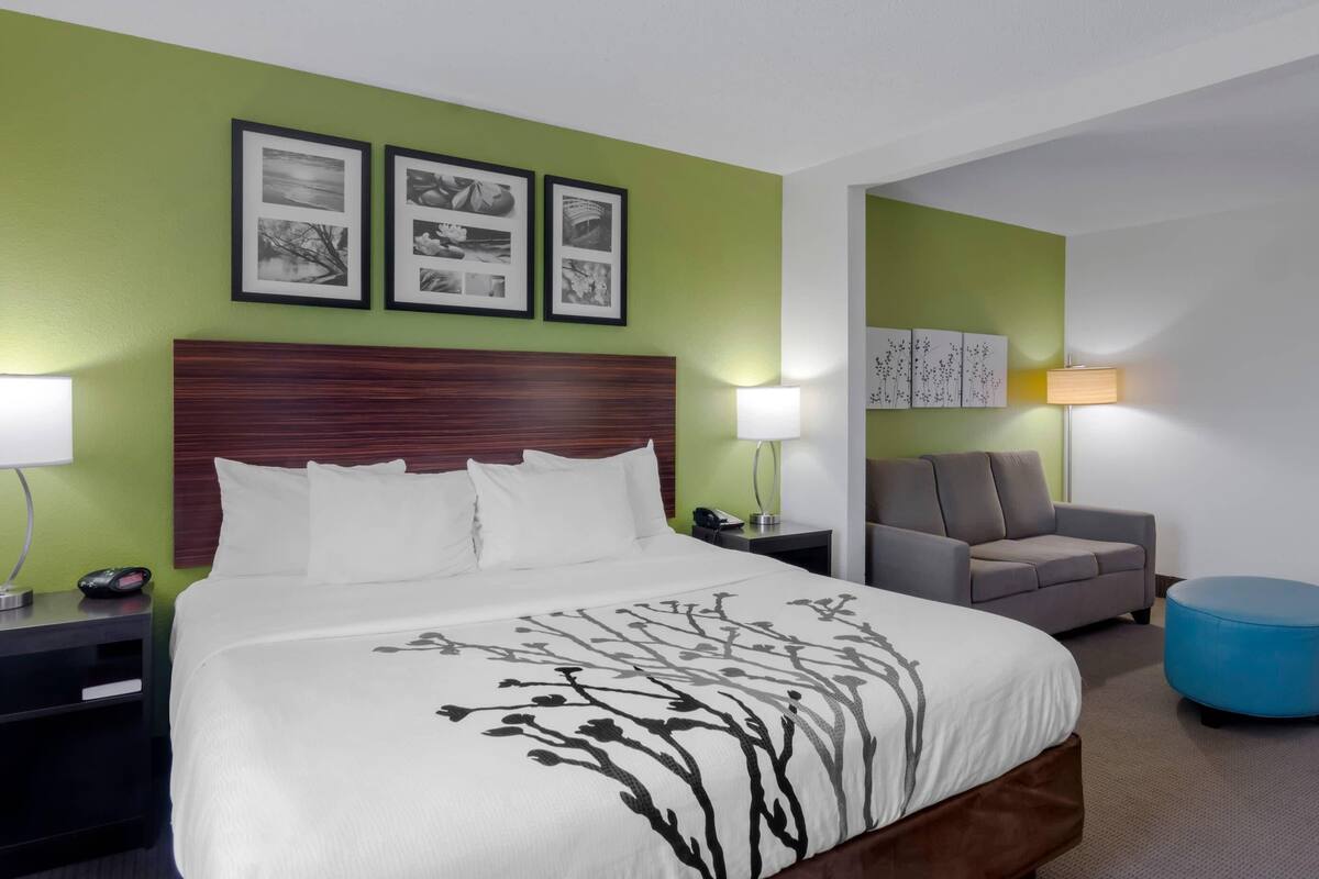 A hotel room with a large bed, a grey sofa, a blue ottoman, and a grey armchair. There are framed pictures on the wall and a lamp on the nightstand.
