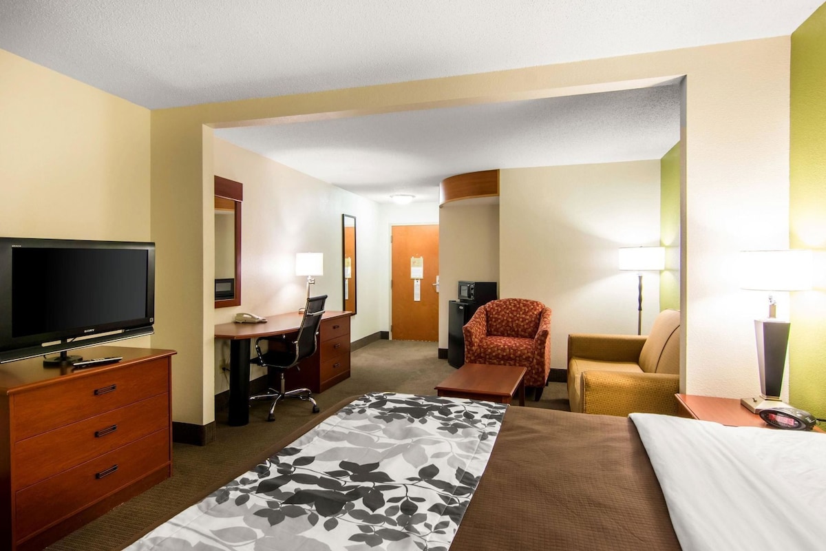 A hotel room with a bed, a desk with a chair, a television, a lamp, and a small table.