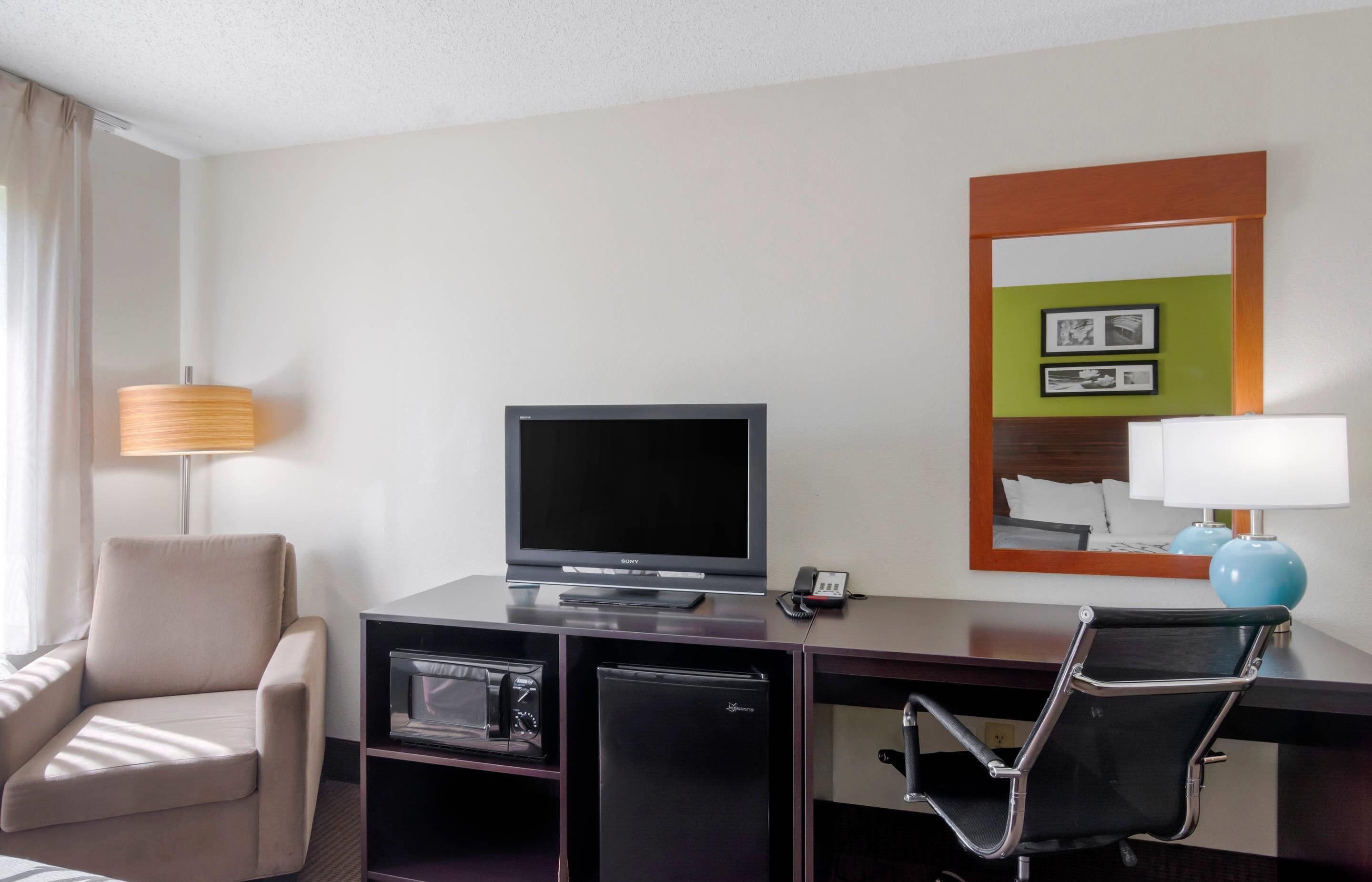 A hotel room with a flat-screen TV, a desk with a lamp, a chair, a minibar, and a small refrigerator.