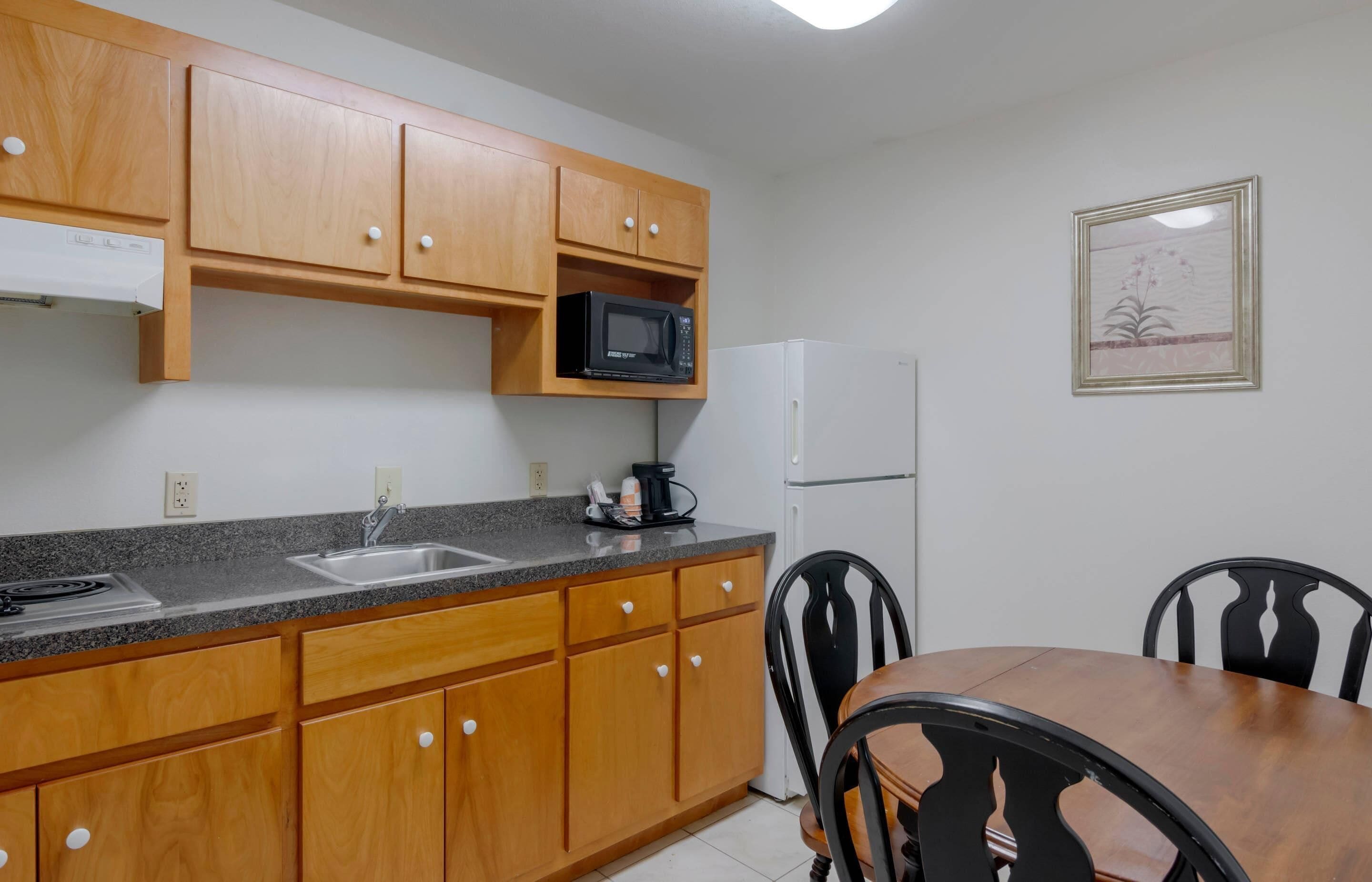 A compact kitchen with wooden cabinets, a granite countertop, a microwave, a coffee maker, a sink, a refrigerator, and a dining table with chairs.