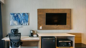 A modern hotel room with a desk, chair, coffee maker, microwave, and a flat-screen TV mounted on the wall.