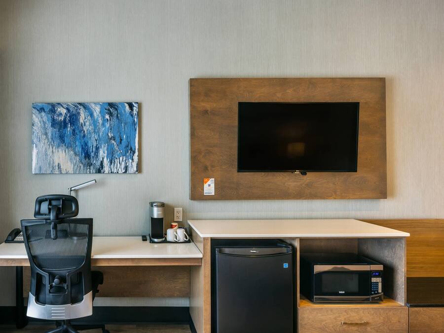 A modern hotel room with a desk, chair, coffee maker, microwave, and a flat-screen TV mounted on the wall.
